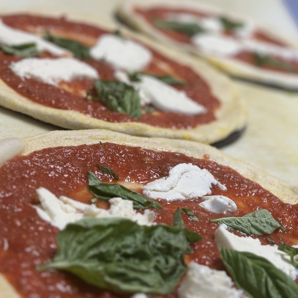 Three uncooked pizzas with red sauce, fresh mozzarella, and basil leaves on a light surface.
