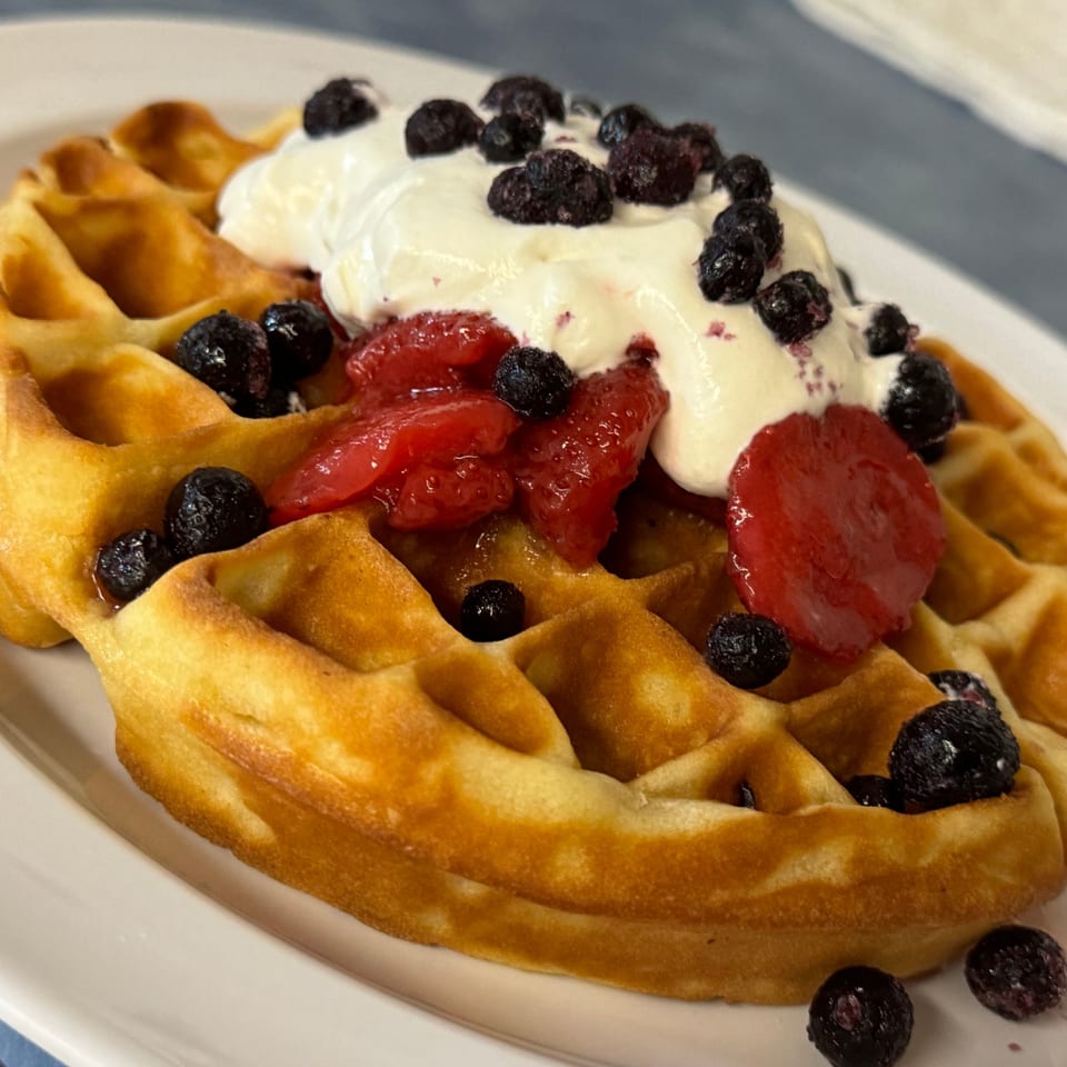 Golden waffle topped with whipped cream, fresh blueberries, and sliced strawberries on a white plate.